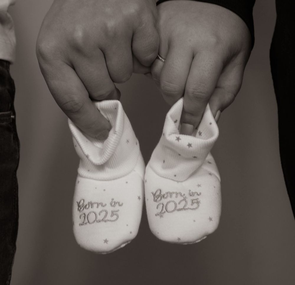 Two hands holding tiny baby booties embroidered with Born in 2025 in black and white.