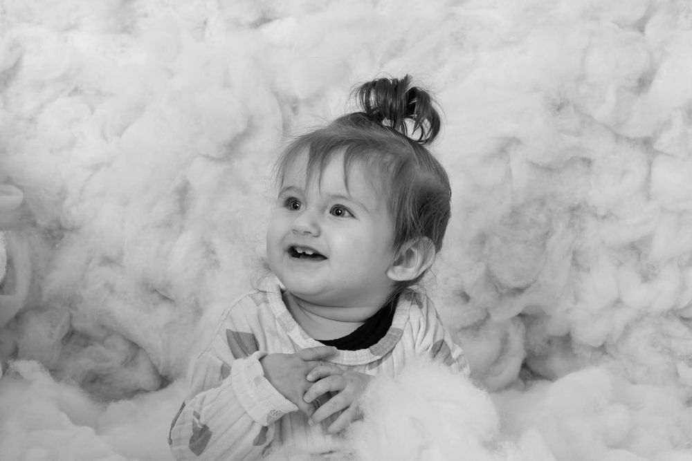 Black and white photo of a smiling baby girl sitting among fluffy white cotton clouds.
