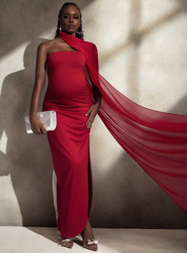 Pregnant woman in red one-shoulder cape gown with slit, holding white clutch, wearing feathered heels.