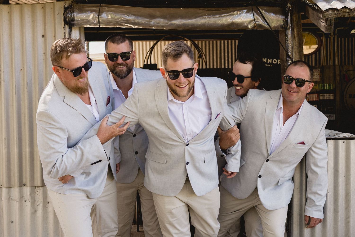 Five groomsmen in matching light beige suits and sunglasses posing together outdoors at a wedding celebration.