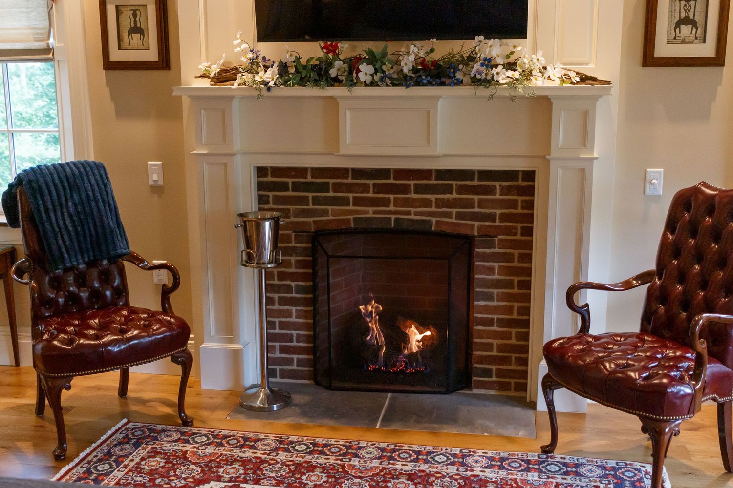 Cozy brick fireplace with white mantel, garland decor, and two red leather tufted chairs on a Persian rug.