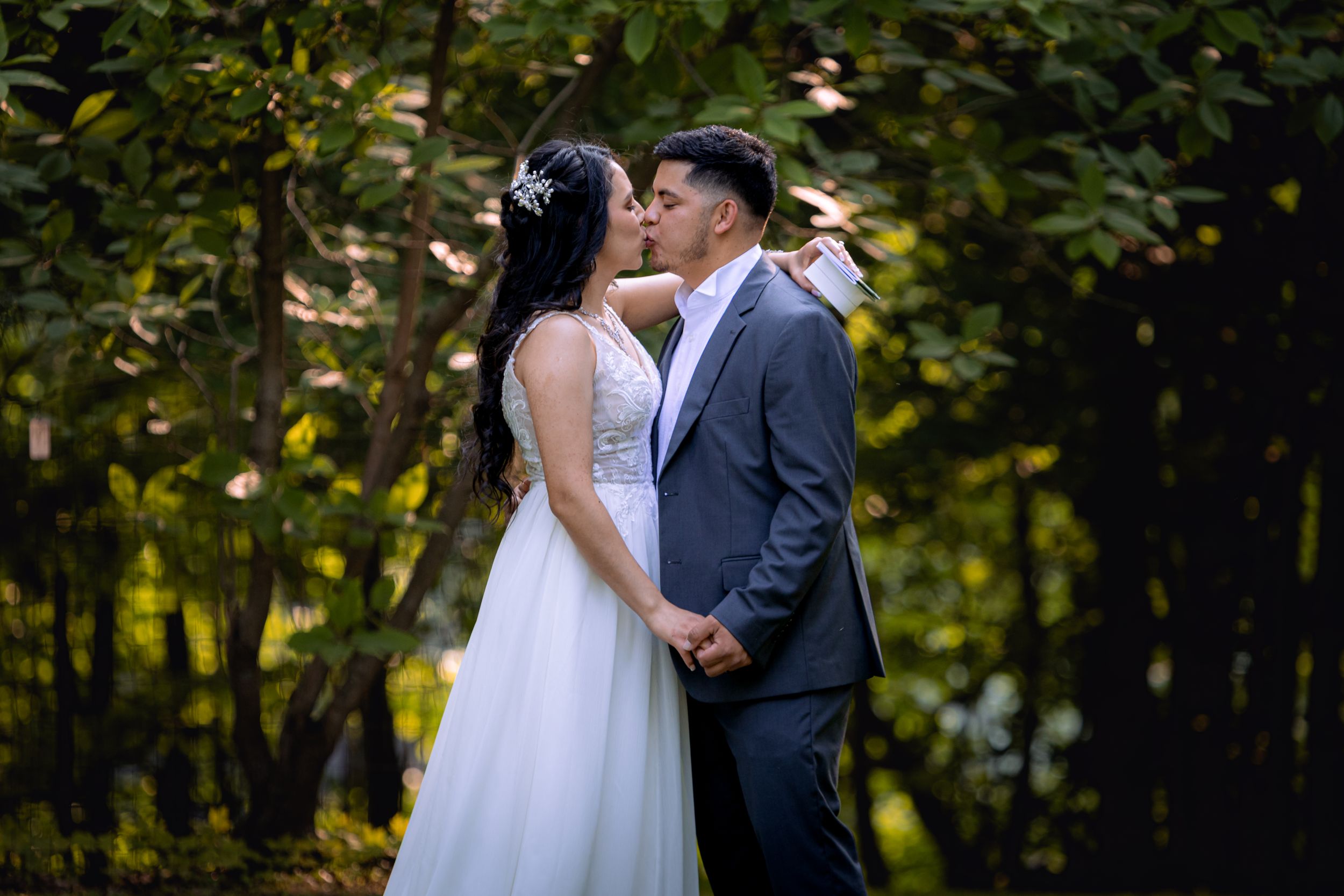 A bride and groom share a romantic kiss surrounded by lush green trees on their wedding day.