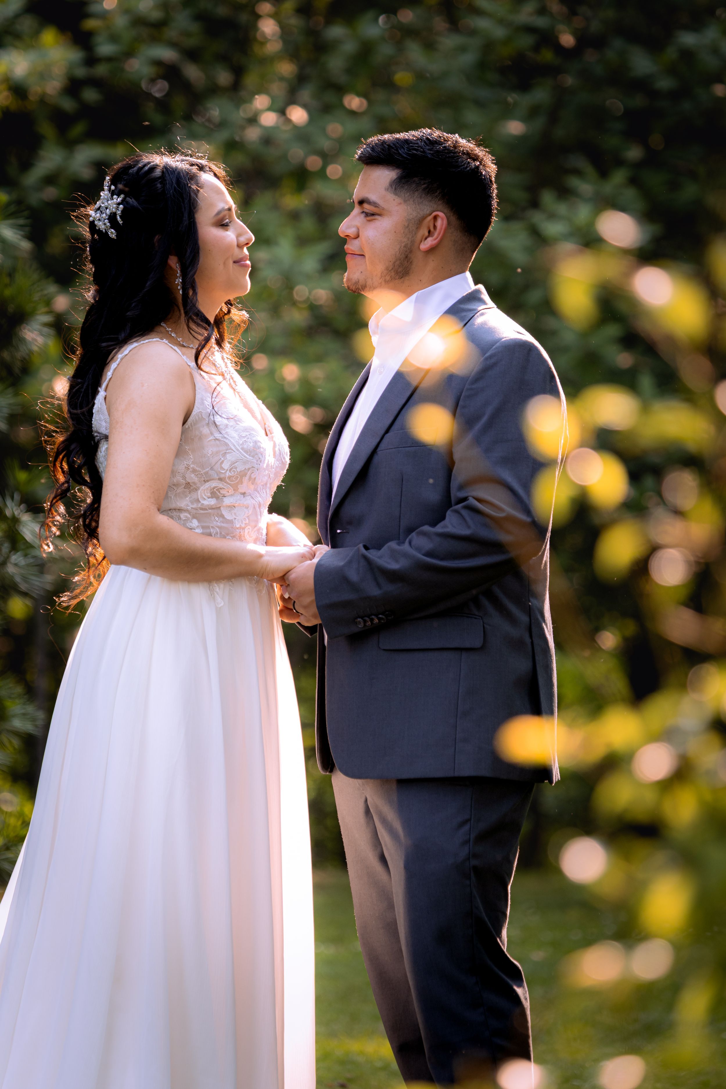 Bride and groom holding hands outdoors in golden sunlight, surrounded by lush green foliage during wedding ceremony.