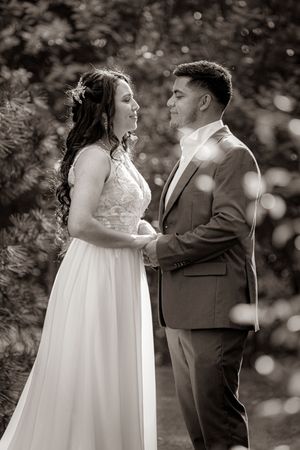 Black and white photo of a bride and groom holding hands and smiling at each other outdoors among lush greenery.