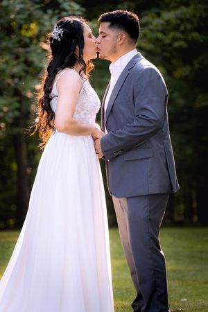 Bride and groom sharing a kiss outdoors, wearing white gown and gray suit, surrounded by lush green trees.