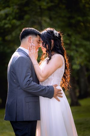 Bride and groom share a romantic forehead touch outdoors, she in a white gown, he in a gray suit, surrounded by lush greenery.