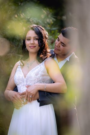 A bride in a white lace gown and groom in a suit embrace outdoors, surrounded by lush greenery, in a romantic wedding portrait.