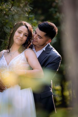 A bride and groom share a tender embrace outdoors, surrounded by lush green trees on their wedding day.