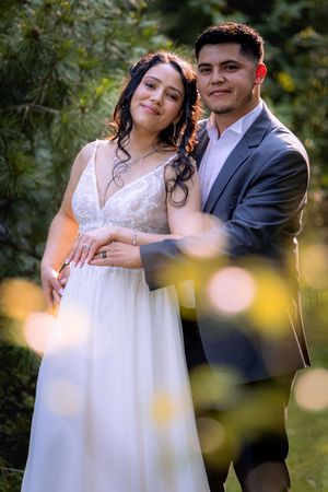 A bride and groom pose together in a lush garden, smiling warmly in wedding attire with soft bokeh lights in the foreground.