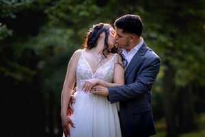 A bride and groom share a romantic kiss outdoors surrounded by lush green trees in a stunning wedding portrait.