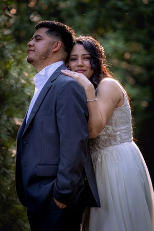 Bride in white lace gown rests her head on groom's shoulder in a romantic outdoor wedding portrait.
