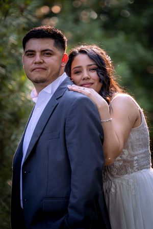 Happy couple poses together outdoors, bride in lace gown resting on groom's shoulder in gray suit.