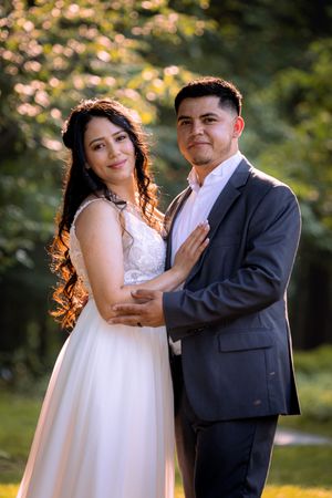 A bride and groom pose together outdoors in golden sunlight, surrounded by lush green trees on their wedding day.