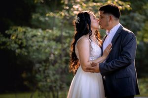 A bride and groom share a romantic kiss outdoors, surrounded by lush green trees in soft golden sunlight.