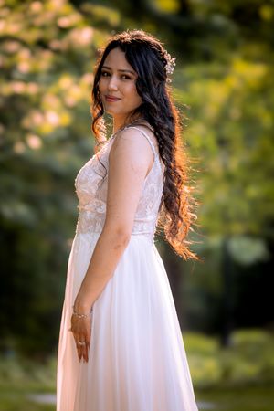 A bride in a white lace gown with long dark hair poses outdoors in a sunlit garden setting.