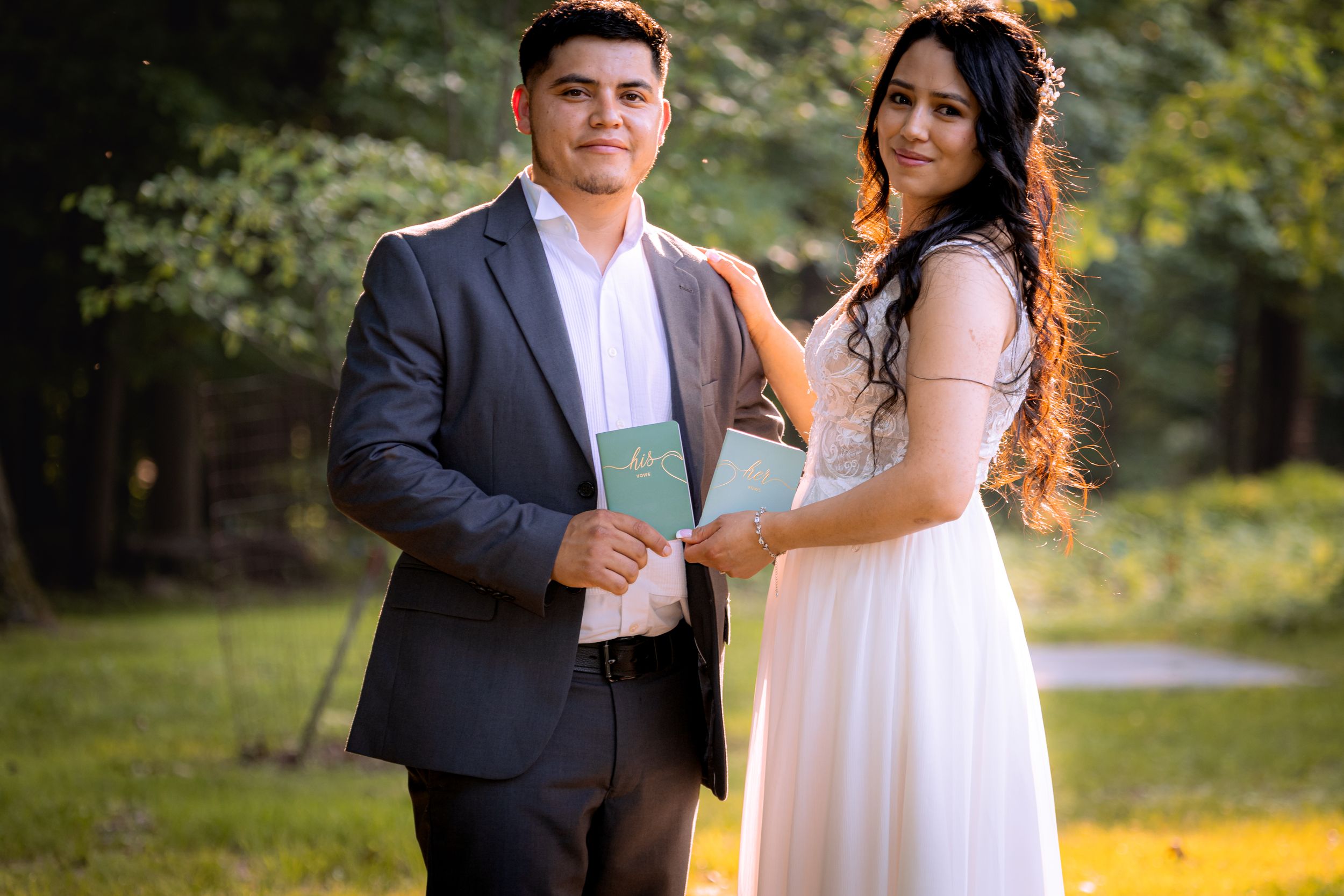 Couple in formal wedding attire holding green books outdoors in a lush green park setting.