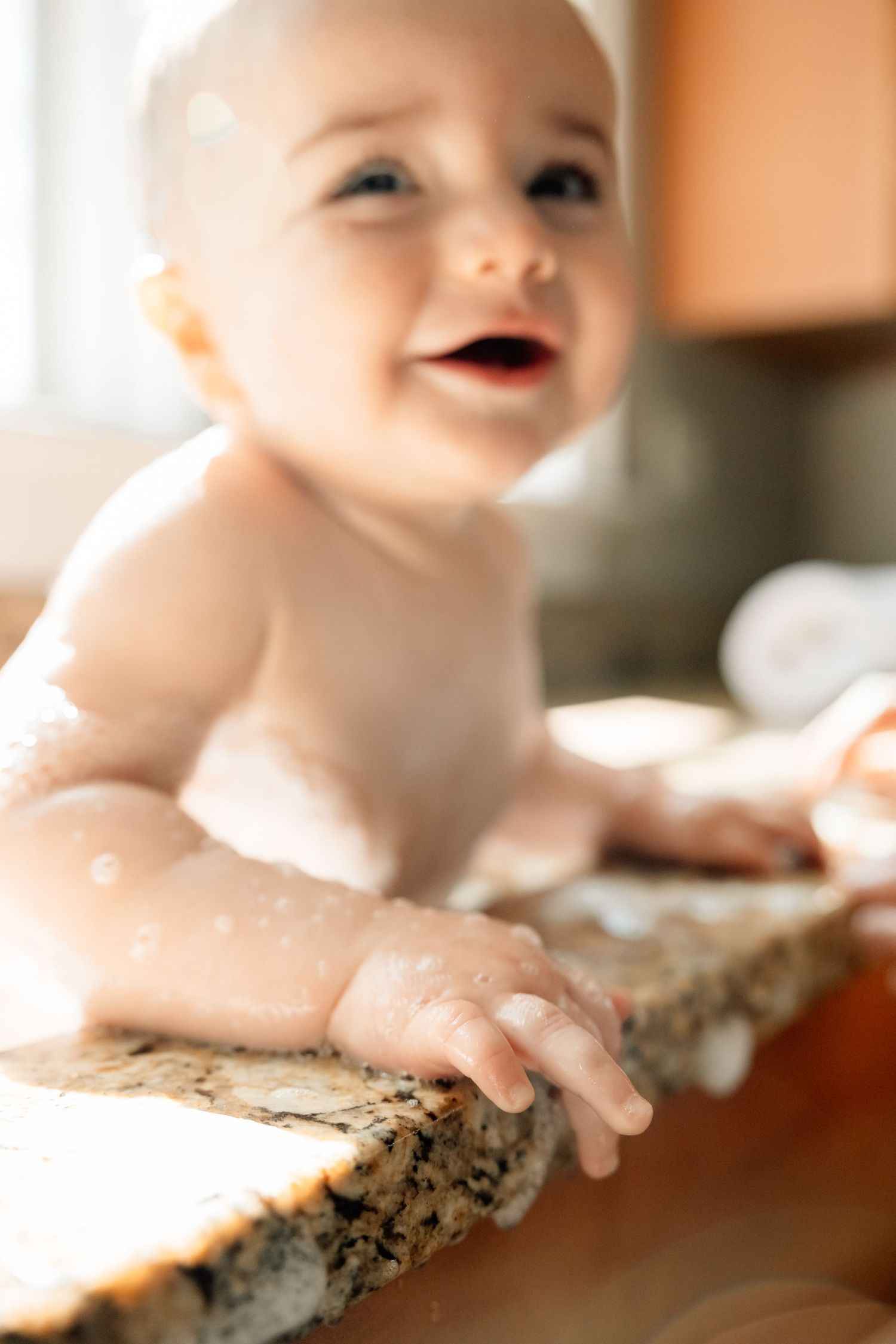 A happy baby covered in soap suds grips the edge of a granite countertop during bath time.