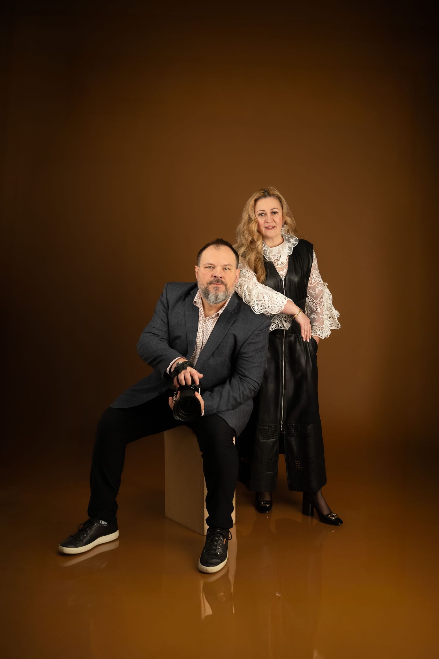 A man and woman pose professionally in a studio against a warm brown background, dressed in elegant dark attire.