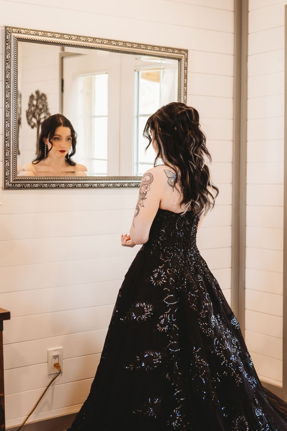 Woman in black sequined ball gown with tattoos views her reflection in ornate mirror.