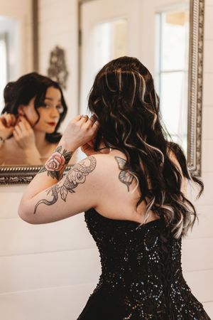 Woman in black sequin strapless gown with floral tattoos adjusts hair while gazing into ornate mirror.