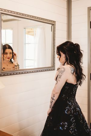 Tattooed woman in black sequin gown admires her reflection in ornate silver mirror against white shiplap wall.