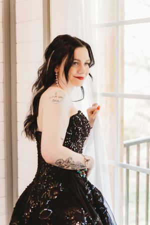 Bride in black sequin strapless gown with tattoos and veil poses by window with red lips and dark hair.
