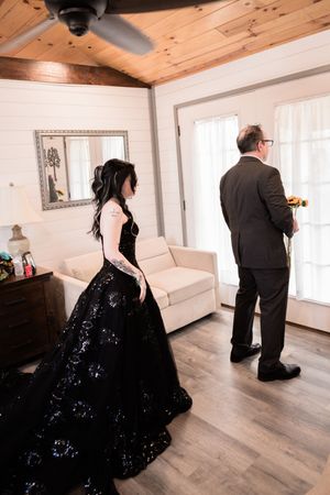 Bride in black sequin ball gown stands near groom holding flowers during first look in rustic white wood-paneled room.