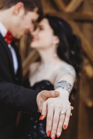 Couple sharing a romantic moment displaying an engagement ring, bride with tattoos and red nails wearing a dark dress.