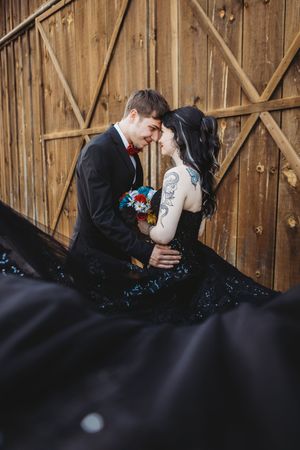 Couple in gothic wedding attire embrace near wooden barn doors, bride in black sequin gown with tattoos holding colorful bouquet.