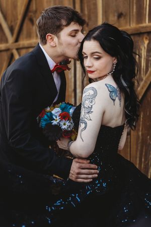 Groom in black suit kisses tattooed bride in black sequin gown holding colorful bouquet near wooden wall.