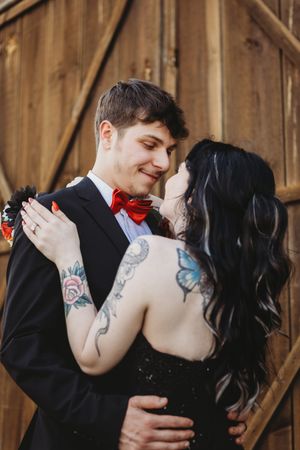 Tattooed couple in wedding attire embracing, groom in black suit with red bow tie, bride in black dress.