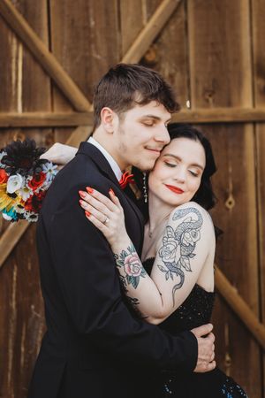 Couple embracing in front of rustic wooden barn doors, bride in black dress with tattoos holding groom in suit with red bow tie.