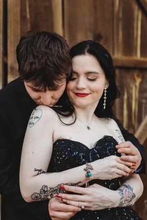 A couple embraces romantically, woman in black sequin dress with tattoos and red lips, man in black suit kissing her shoulder.