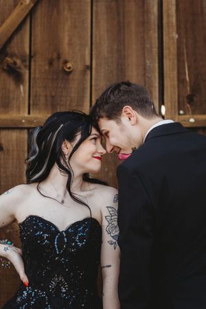 A couple in formal attire share a romantic moment, the woman in a black sequin gown with tattoos, against a wooden backdrop.