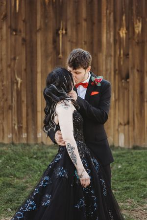 A couple shares a romantic kiss, bride in black embroidered gown with tattoos, groom in black suit with red bow tie.