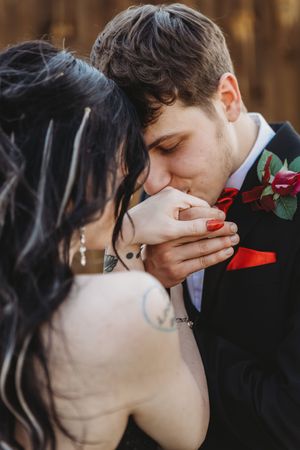 Groom in black suit kissing bride's hand at wedding, red rose boutonniere, bride with dark hair and tattoo.
