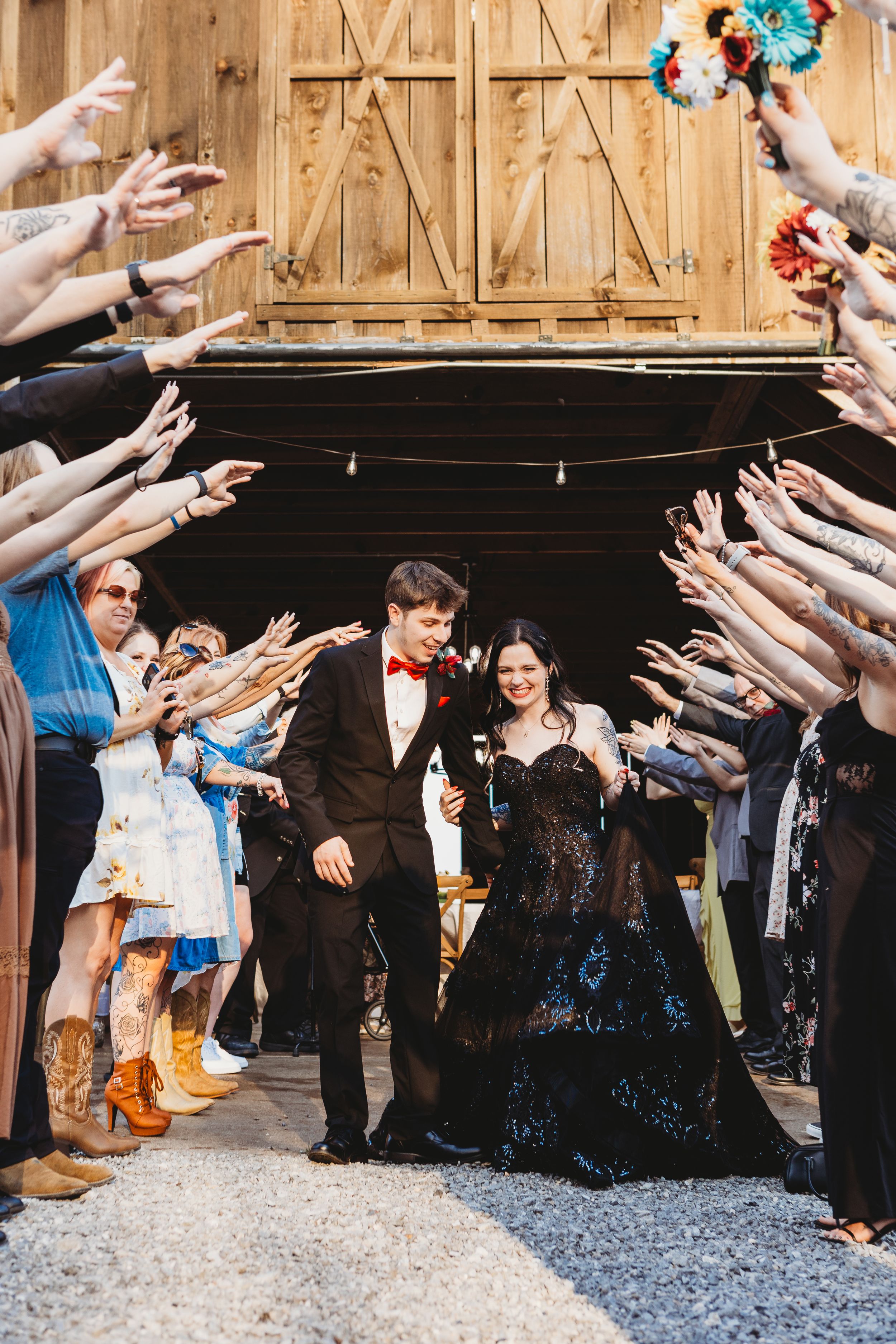 Bride in black lace gown and groom in tuxedo exit barn wedding venue through tunnel of guests reaching hands.