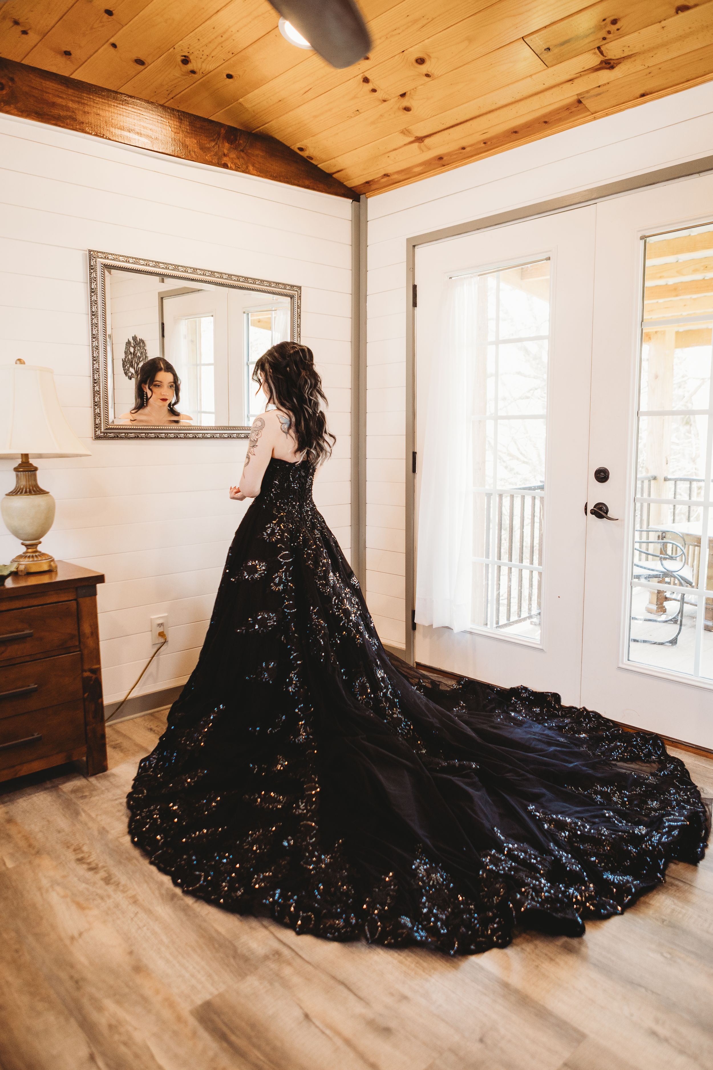 Woman in stunning black sequined ballgown with long train stands in rustic room, reflected in mirror.