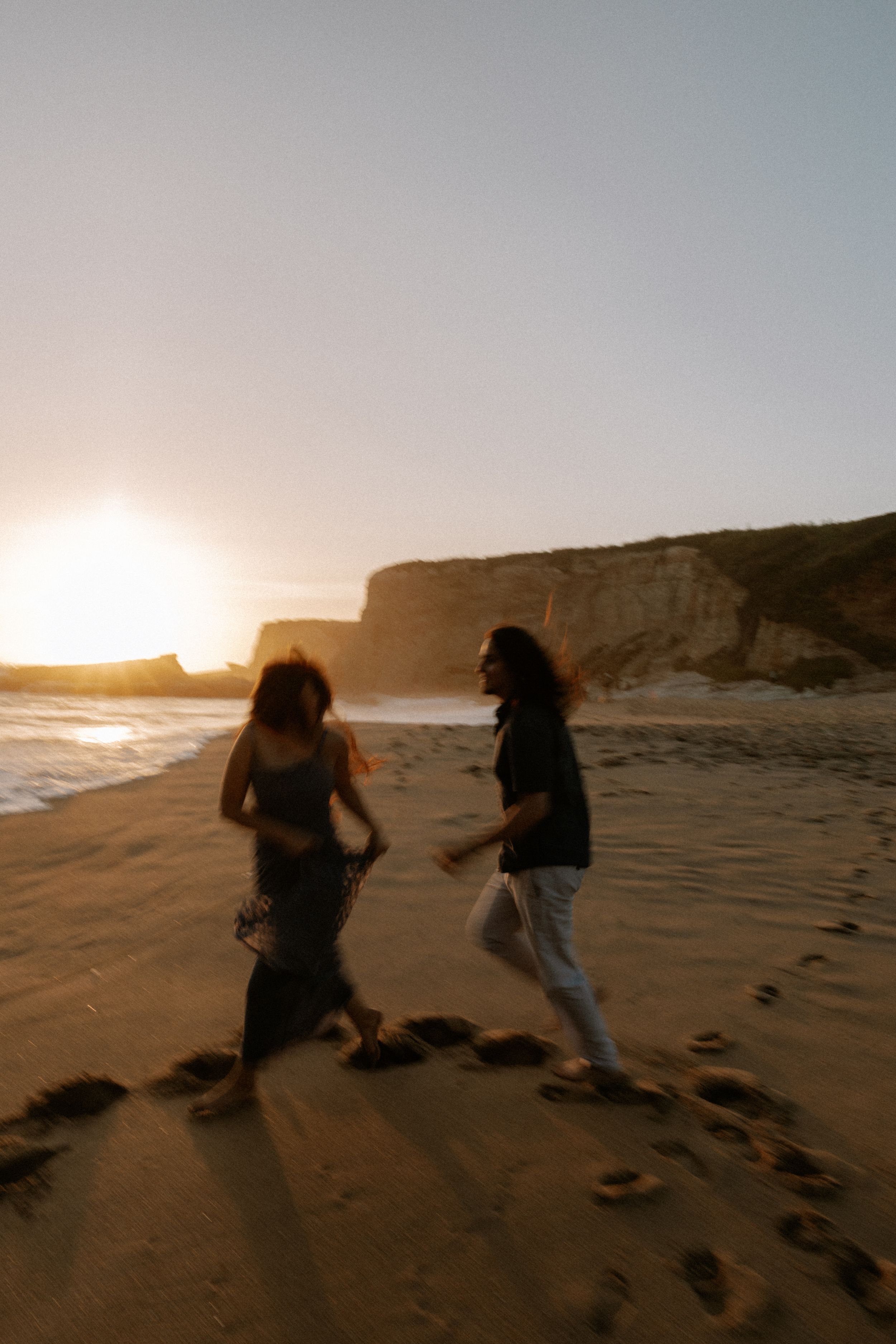 Two people running playfully on a sandy beach during a golden sunset by coastal cliffs.