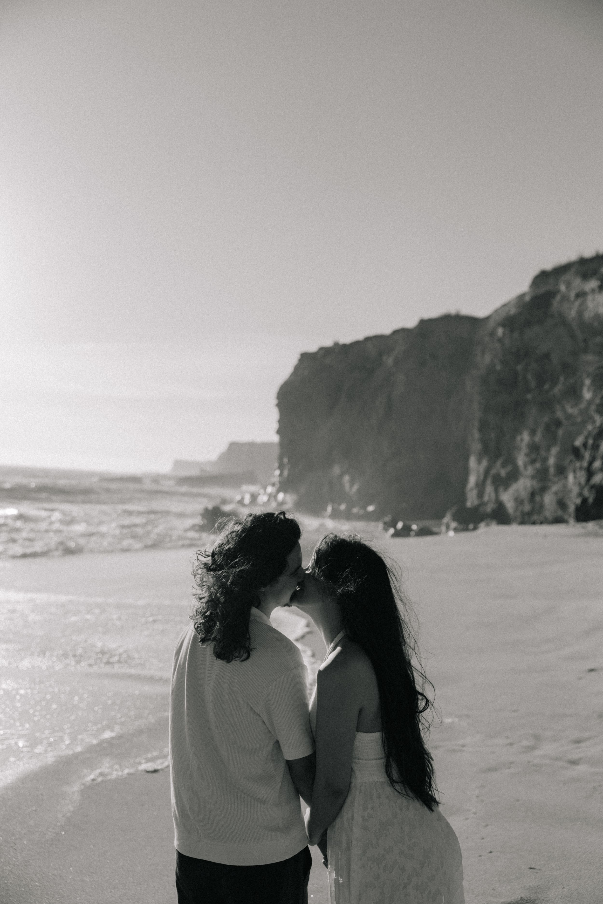 Black and white photo of a couple sharing a kiss on a sandy beach with dramatic cliffs in the background.