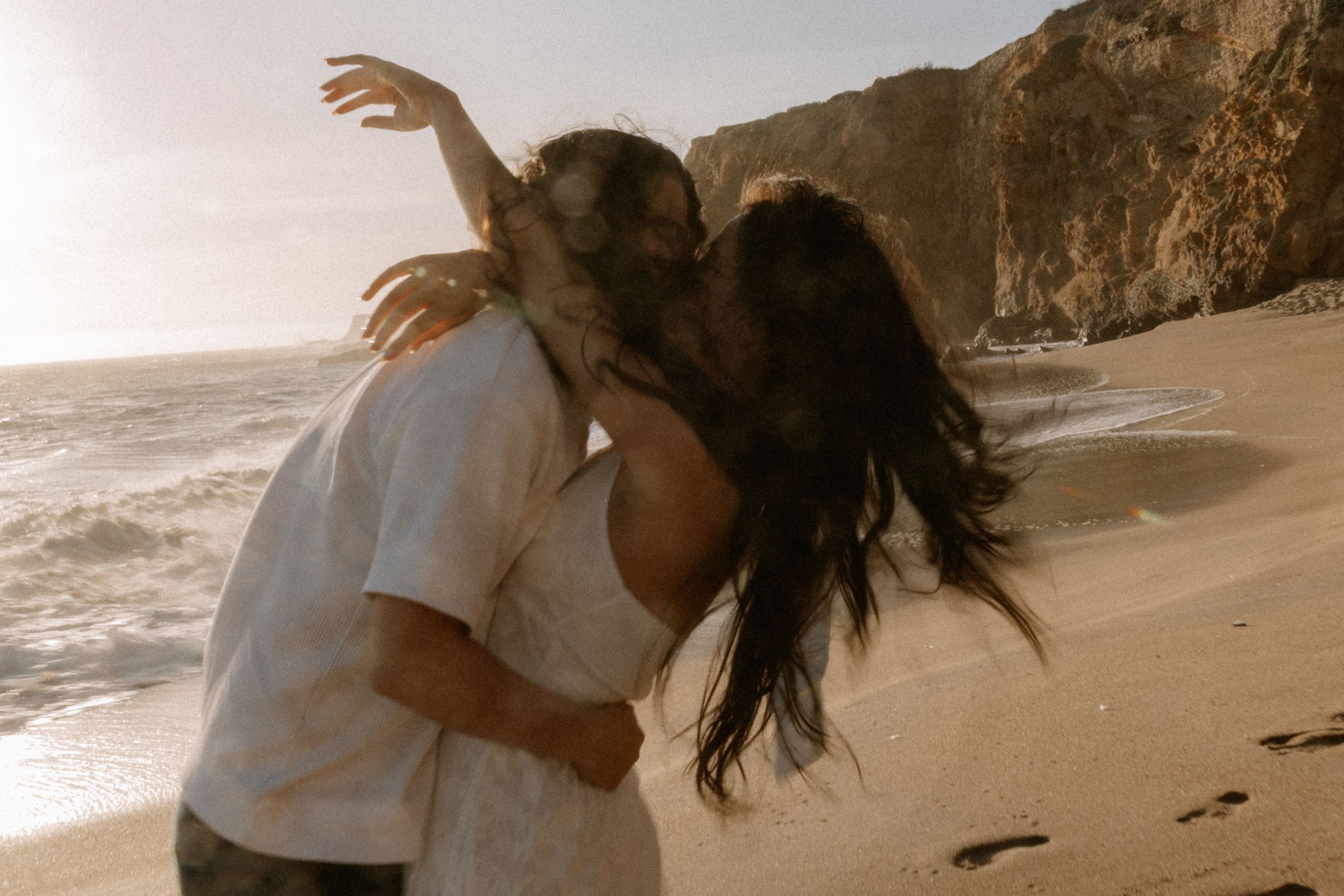 Couple sharing a romantic kiss on a scenic beach with dramatic cliffs and ocean waves in golden sunlight.