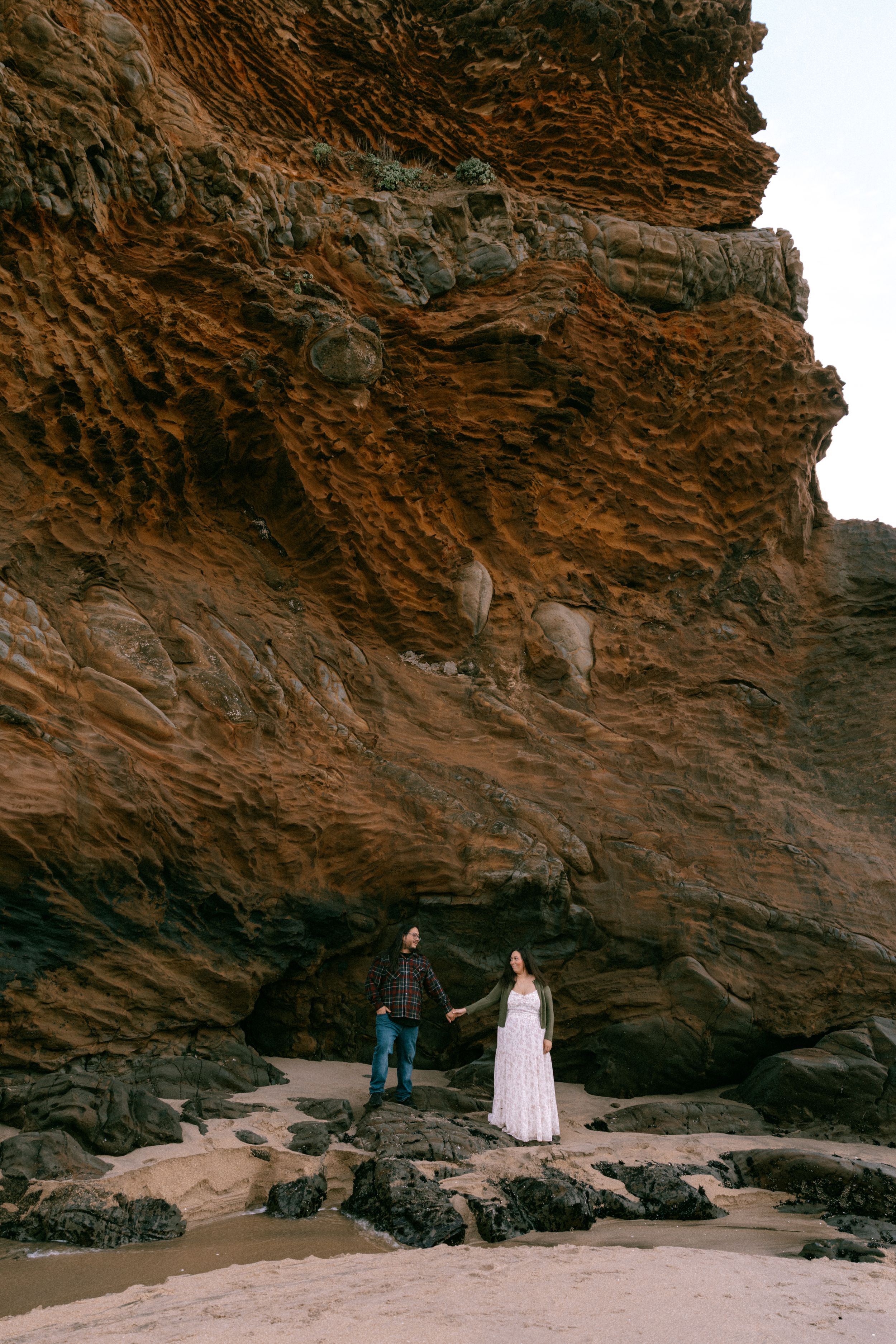Couple holding hands on rocky beach beneath dramatic orange cliff formations.