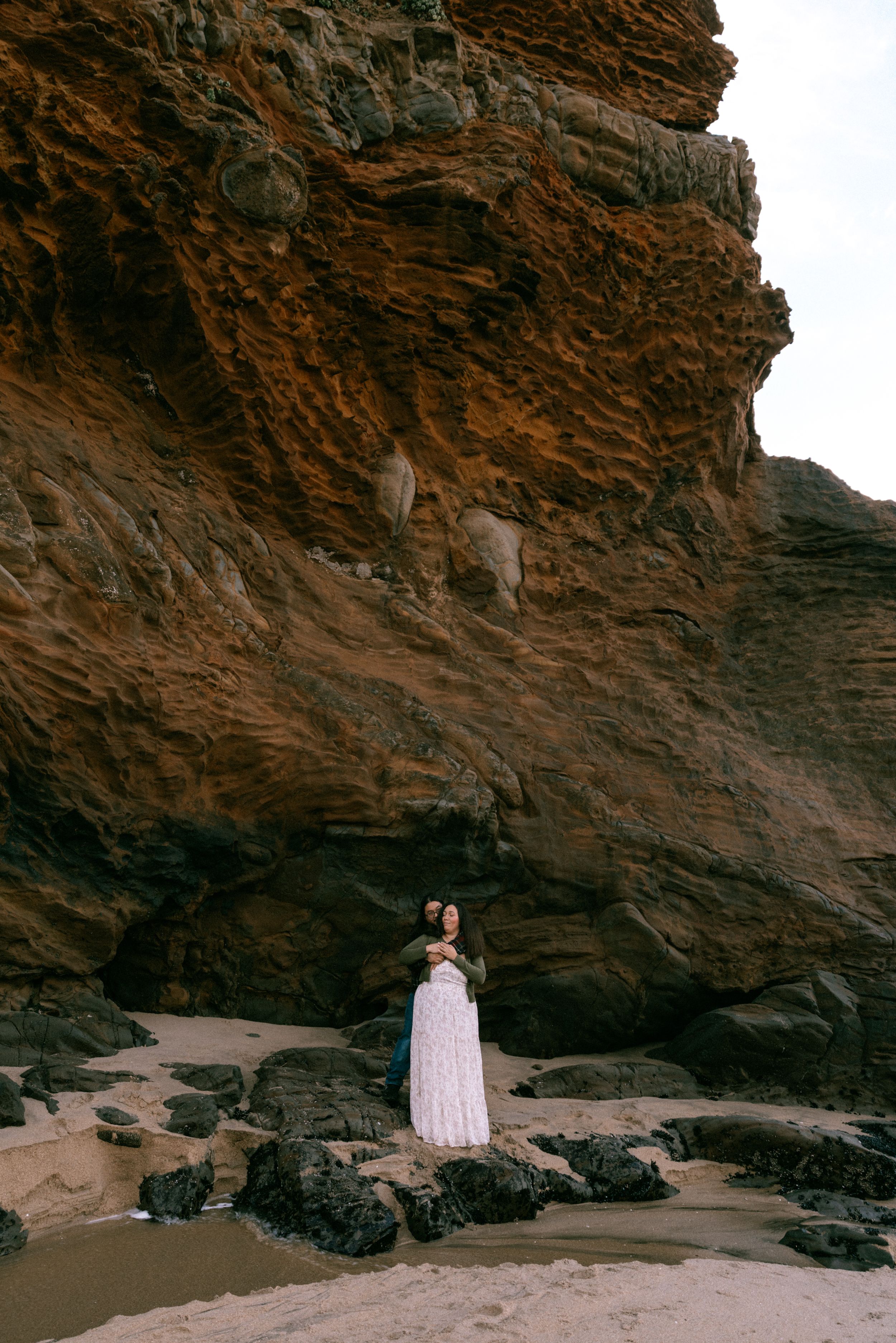 Couple embracing on rocky beach beneath dramatic red sandstone cliffs, woman in flowing pink dress, romantic outdoor portrait.
