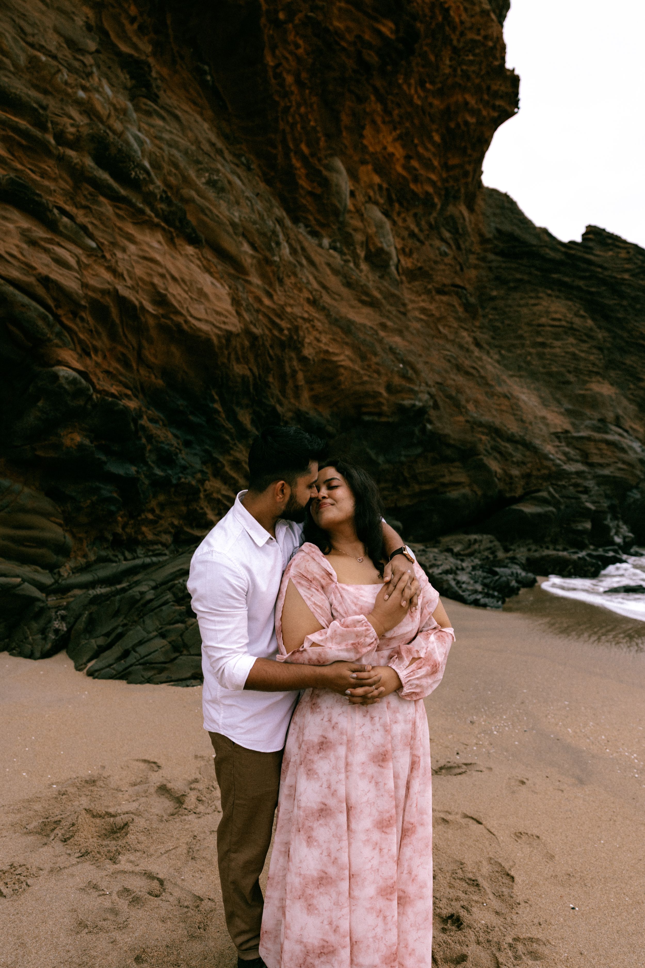 Couple embracing romantically on a sandy beach with dramatic rocky cliffs backdrop.