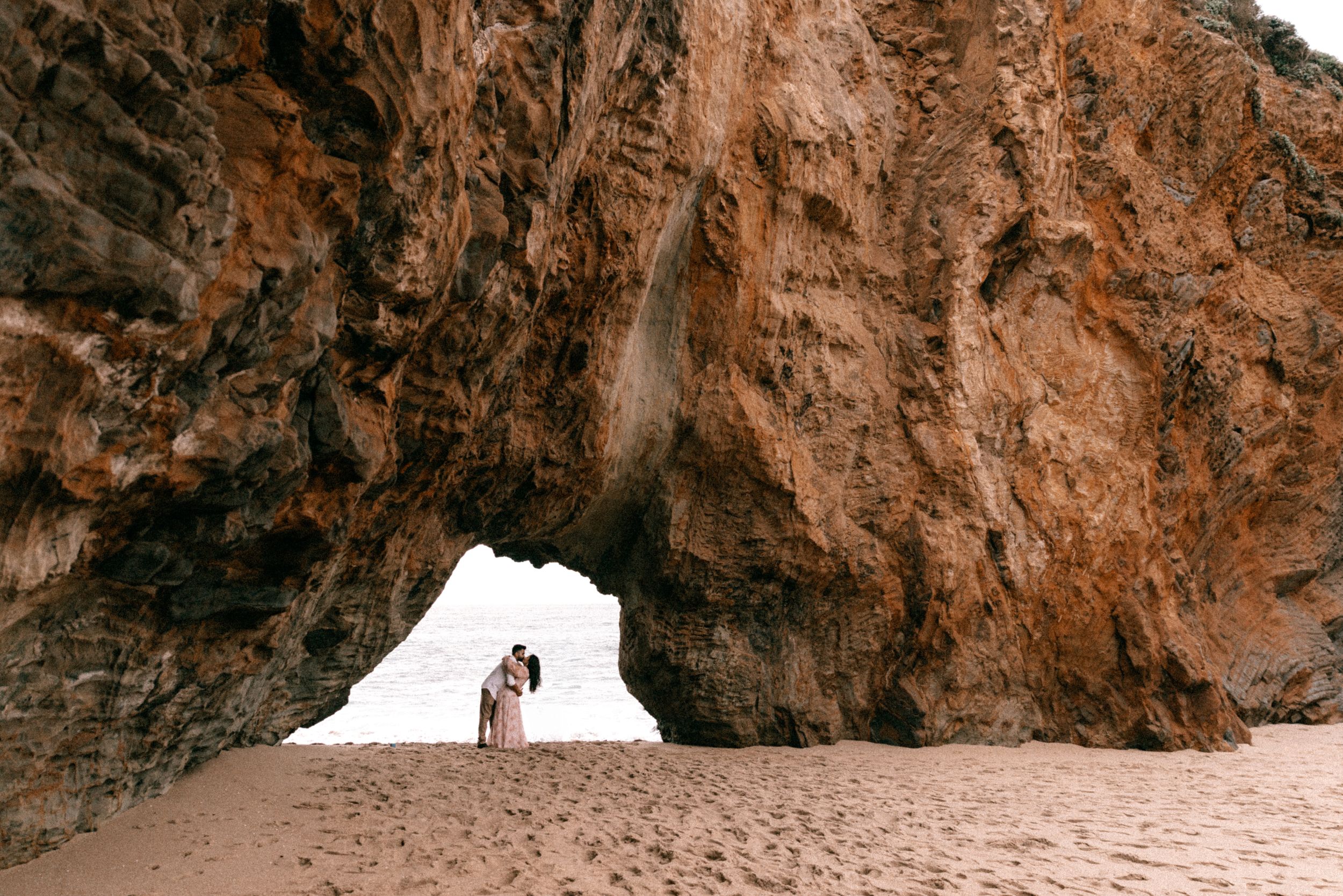 Couple sharing a romantic kiss under a dramatic natural rock arch on a sandy beach.