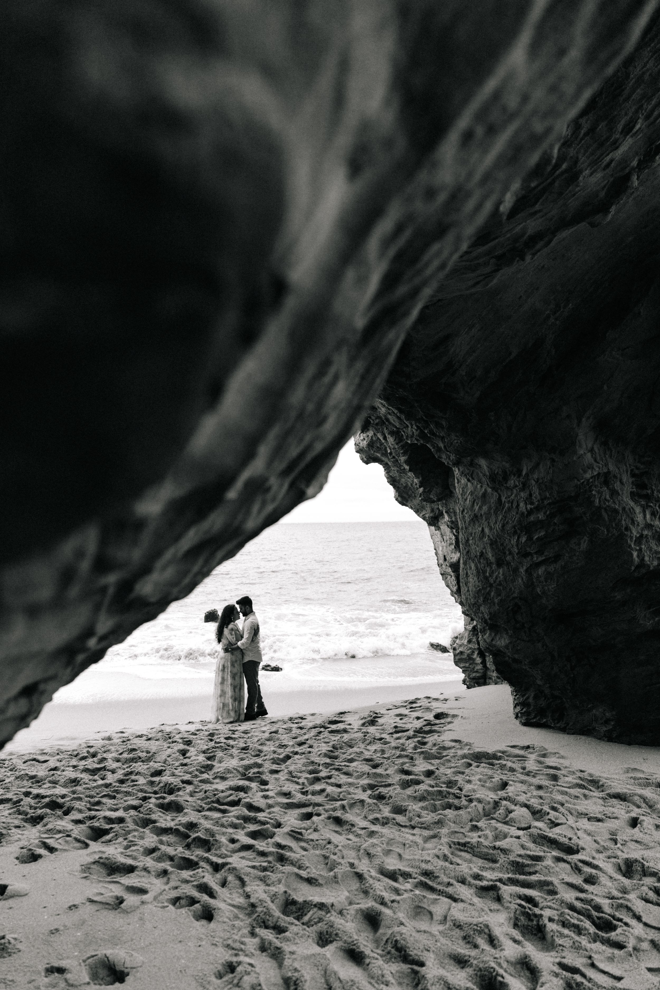Black and white photo of a couple embracing on a sandy beach, framed by a dramatic rocky cave arch with ocean waves.