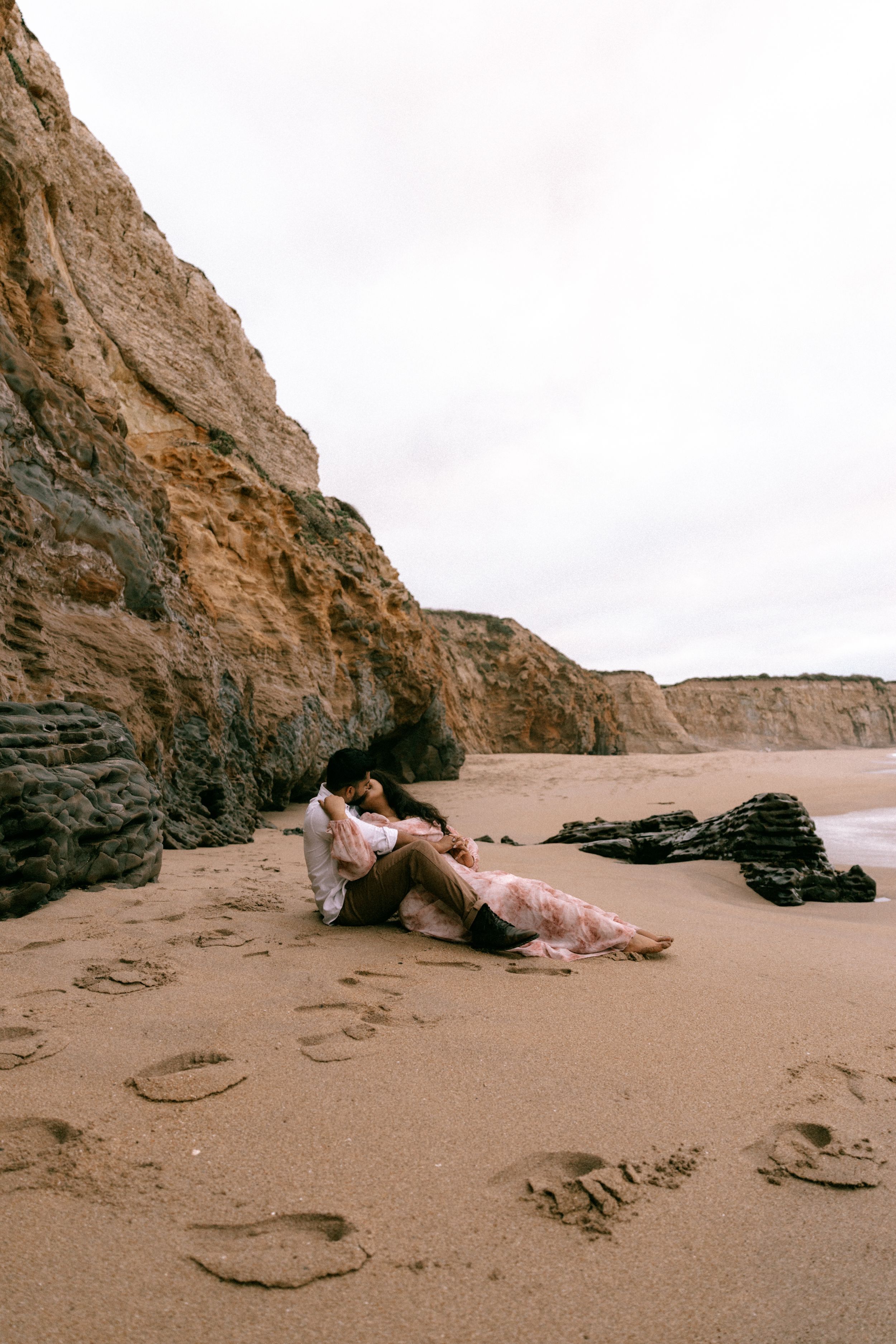 Couple sitting together on sandy beach leaning against towering rocky cliffs.