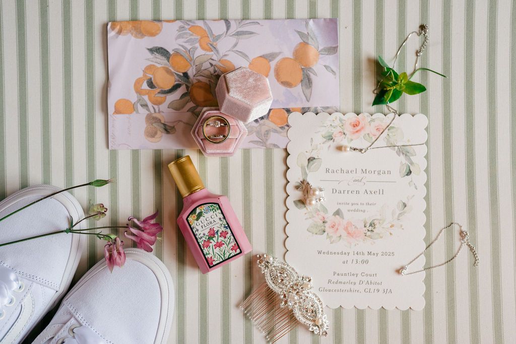 Elegant wedding flat lay with floral invitation, white sneakers, pink perfume bottle, and delicate hair accessories on striped background.