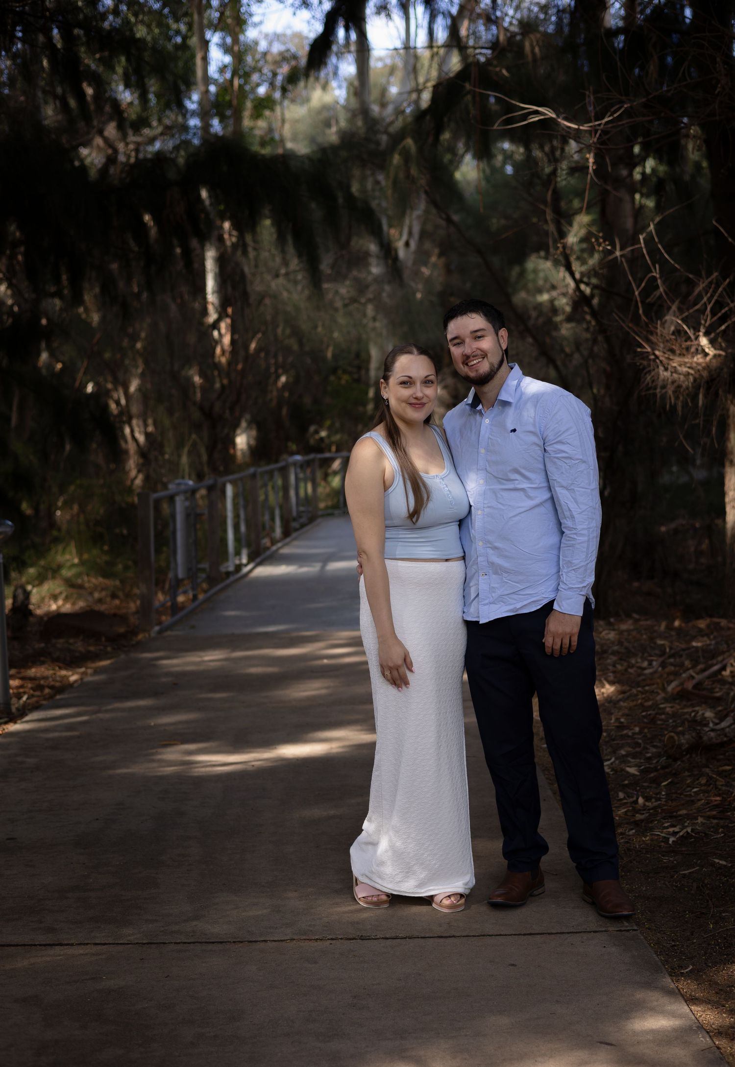 Couple posing on a tree-lined pathway, woman in white dress and man in blue shirt and navy pants.