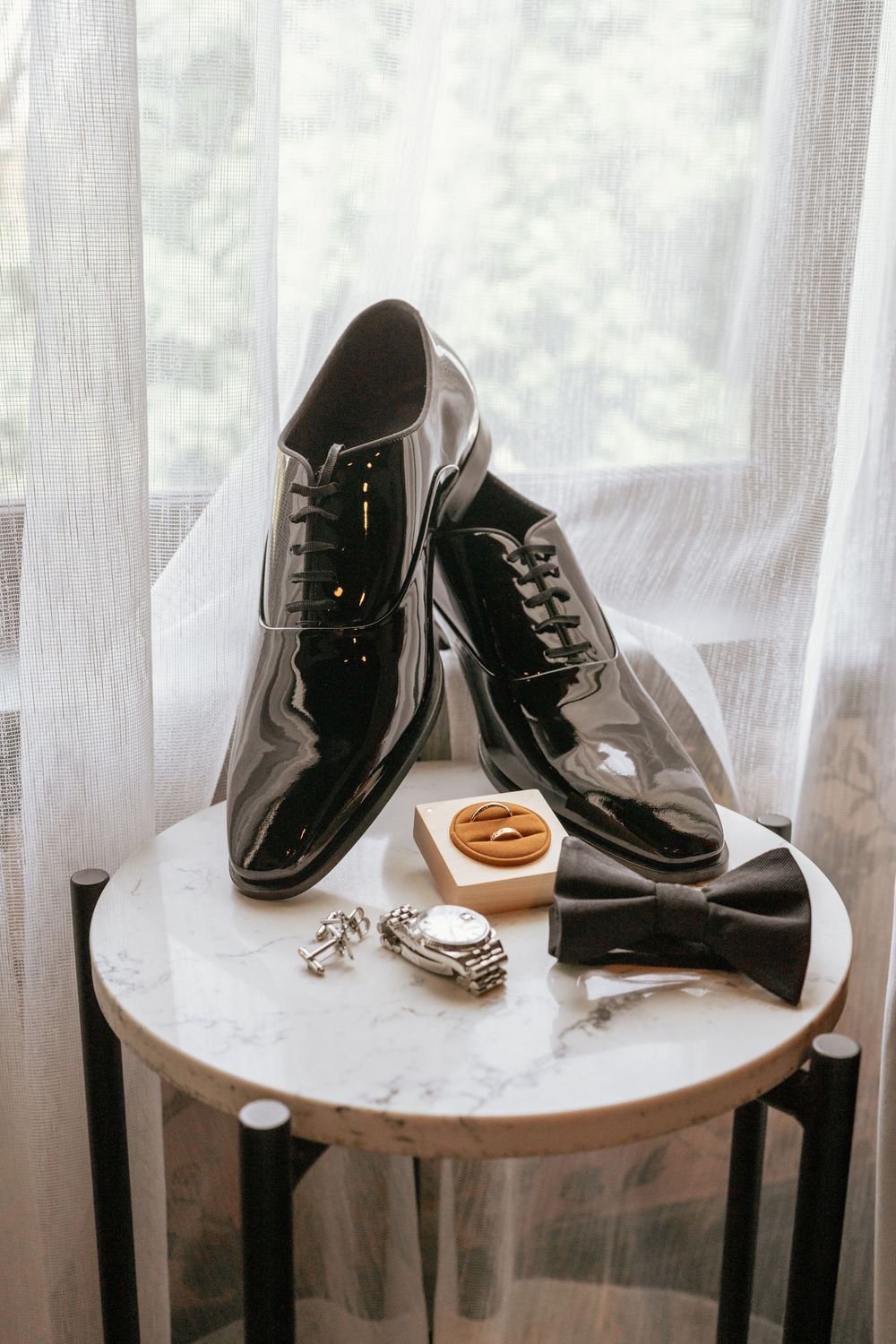 Groom wedding accessories display with black patent leather oxford shoes, bow tie, watch, cufflinks, and wooden ring box on marble table.